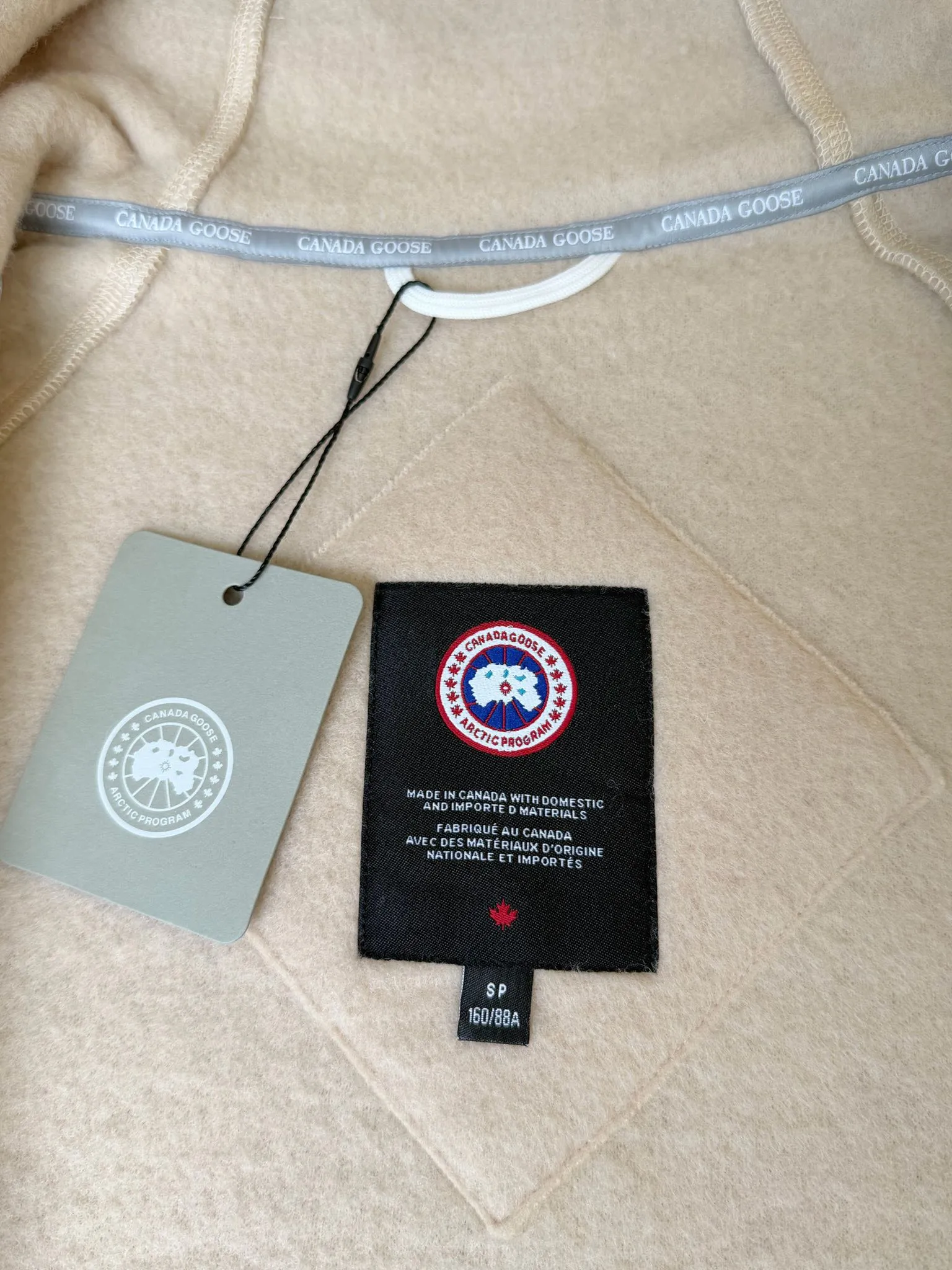 P660 Canada Goose Fleece Wool Badge Hooded Jacket Fabric selection: Wool fleece fabric blended with wool as the main body and a very small amount of spandex and polyester fiber. Wool is a core natural ingredient that provides unparalleled temperature regulation, antibacterial and anti-odor properties and a skin-friendly feel. The feeding of a small amount of spandex gives the fabric a certain degree of four-way elasticity, making it non-restrictive during daily activities. As the basis of fleece's structure, polyester fiber enhances the fabric's durability, wrinkle resistance and quick-drying properties. It contributes to the loft of the fleece and provides functions such as trapping air.  Detail 1: The sleeves and hem are closed with 2×2 ribbing. Its strong elasticity can closely fit wrists and waists of different thicknesses, forming an effective 'seal' to prevent cold wind from blowing back. It can still maintain its original shape after repeated stretching and use, and is not easy to loose and deform. And the ribbed cuffs can convey a sense of design and enhance the visual beauty.  Detail 2: The combination of hood design and elastic buckle improves warmth and windproof performance when wearing. Accessories: Customized elastic drawstring, elastic buckle, custom zipper pendant, custom armband Size: S M L Color: black gray coffee rice pink